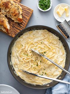 a pan filled with pasta and chicken on top of a wooden cutting board next to bread