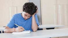 a young man sitting at a desk writing on a notepad with a pen in his hand