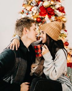 a man and woman sitting in front of a christmas tree with their noses to each other