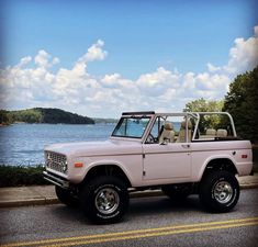 an old pink truck parked on the side of the road next to some water and trees