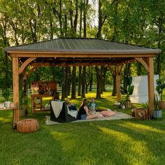 two people sitting under a wooden gazebo in the middle of some grass and trees