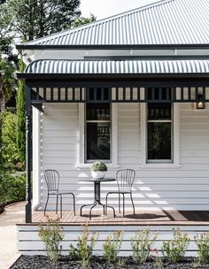 a small white house with two chairs and a table on the front porch, surrounded by greenery