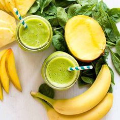 two glasses filled with green smoothie next to sliced mangoes and bananas on a table