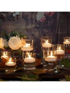 there are many lit candles in glass vases on the table with flowers and greenery