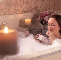 a woman sitting in a bathtub eating something out of a package next to a candle