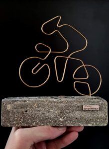 a hand holding a stone block with a wire sculpture on it's end and a bike in the background