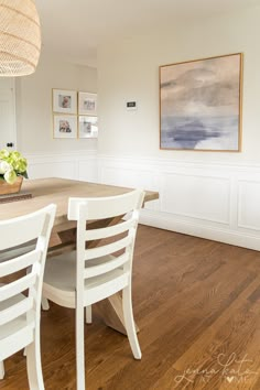 a dining room table with white chairs next to a painting on the wall above it