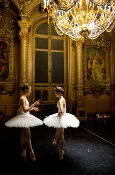 two ballerinas in white tutus standing next to each other with chandelier above them