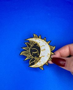 a hand is holding a sun and moon brooch on a blue background with red nail polish