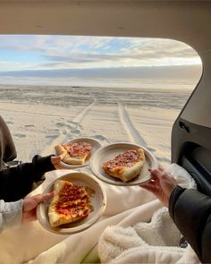 two people holding plates with pizza on them in the back of a car, while another person holds out their hand