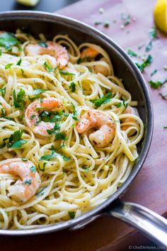 pasta with shrimp and parsley in a pan on a wooden table next to lemon wedges