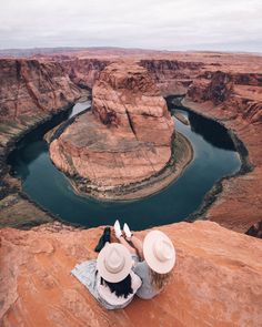 two people sitting on the edge of a cliff looking out over a river and canyon