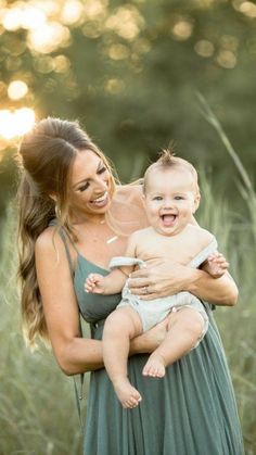 a woman holding a baby in her arms and smiling at the camera while standing in tall grass