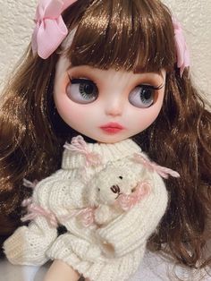 a close up of a doll holding a teddy bear wearing a sweater and pink bow