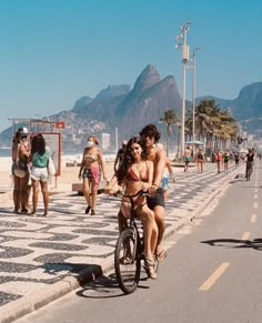 two women riding bikes on the beach with people walking and sitting in the water behind them