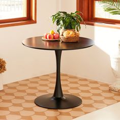 a small table with fruit on it in front of two windows and potted plants