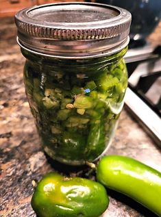 two green peppers sitting on top of a counter next to a jar filled with pickles