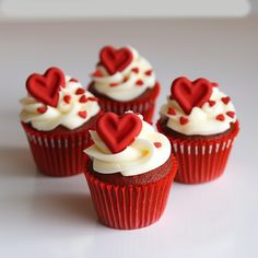 four red cupcakes with white frosting and hearts on top are sitting in a row