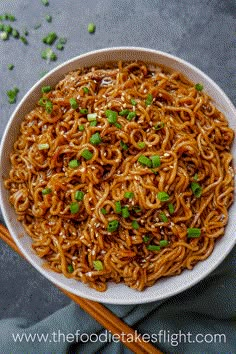 a white bowl filled with noodles and green onions next to chopsticks on a gray surface