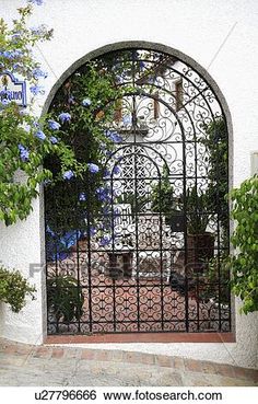 an iron gate in front of a white building with blue flowers growing on the side