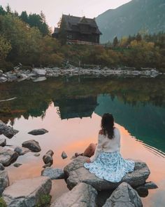 a woman sitting on rocks in front of a body of water with a house in the background