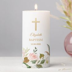 a white candle with a cross on it next to some pink flowers and a vase