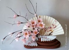 a white fan with pink flowers in it sitting on top of a wooden stand next to a tree branch