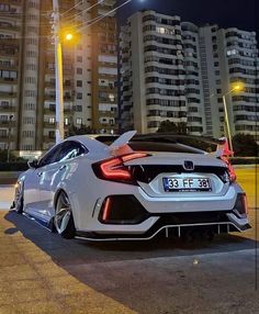 the rear end of a white sports car parked in front of tall buildings at night