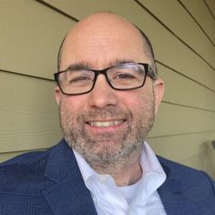 a man wearing glasses and a blue jacket smiles at the camera while standing in front of a house