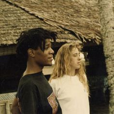 two young people standing next to each other in front of a thatched roof house