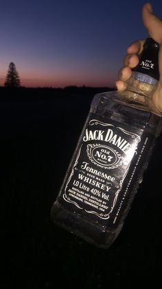 someone holding a bottle of jack daniels whiskey in their hand at night with the sun setting