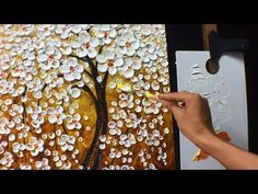 a person is painting a tree with white flowers on yellow and orange background while holding a paintbrush in their left hand