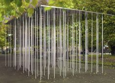 an artistic sculpture in the middle of a park surrounded by trees and grass, with metal rods hanging from it's sides