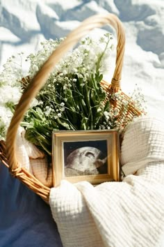 a basket filled with flowers next to a framed photo on a bed covered in blankets