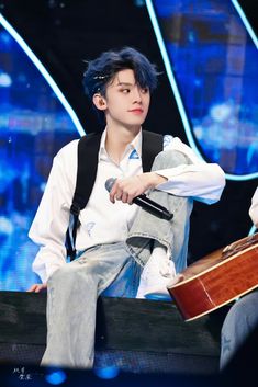 a young man with blue hair sitting on top of a chair holding an acoustic guitar