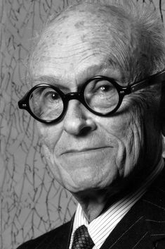 an old man wearing glasses and a suit with writing on the wall behind him in black and white