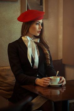 a woman sitting at a table wearing a red hat and holding a coffee cup with a spoon in her hand