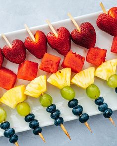 fruit skewers are lined up on a white plate with grapes, strawberries and pineapples