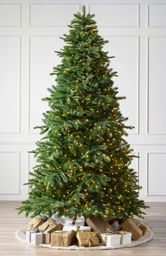 a christmas tree with lights and presents under it on a wooden floor in front of a white wall