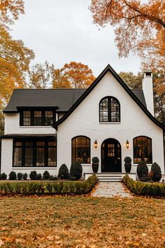 a white house with black roof and windows in the front yard surrounded by fall foliage