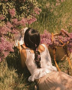 a woman sitting in the grass reading a book next to some flowers and a basket