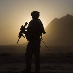 a soldier is silhouetted against the setting sun in an arid area with mountains behind him