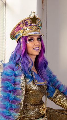 a woman with purple hair wearing a gold and blue dress, crown and feathered jacket