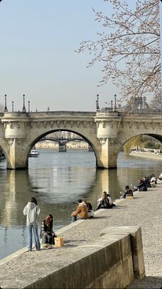 people sitting and standing on the edge of a body of water with an old bridge in the background