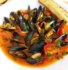 a white plate topped with mussels and tomatoes next to a piece of bread