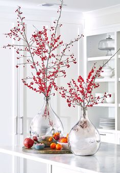 two glass vases with red flowers in them on a counter top next to plates and utensils