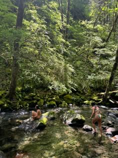 two women in bathing suits are standing on rocks near a stream and some green trees