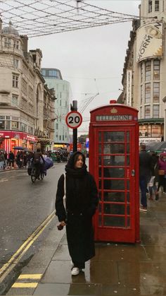 a woman standing next to a red phone booth on the side of a street with people walking by