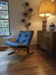 a blue chair sitting next to a lamp on top of a wooden dresser under a window