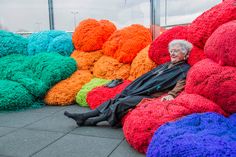 an old woman is sitting on the ground next to colorful knitted balls and pillows